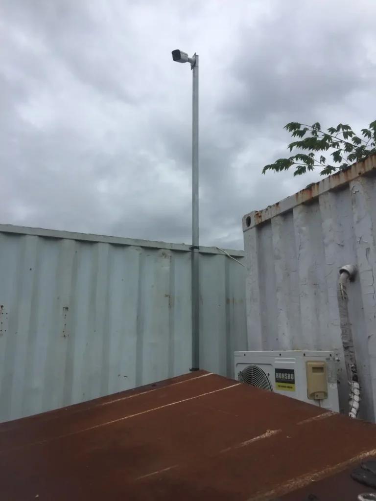 Kamera CCTV Outdoor terpasang kokoh pada tiang besi di samping dinding kantor kontainer (Direksi Keet) proyek konstruksi area Surabaya oleh tim CCTVSurabaya.com.